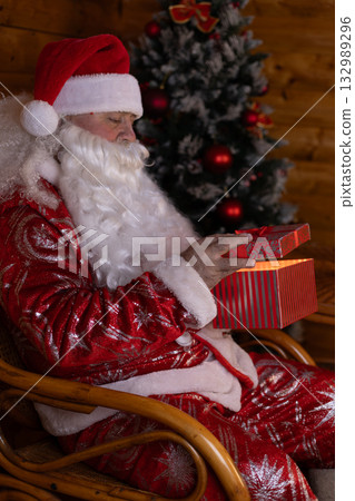 Santa Claus sits in a chair near a Christmas tree, holding a gift box Santa Claus sits in a chair near a Christmas tree, holding a gift box 132989296
