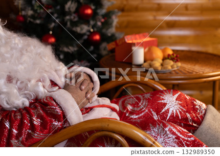 Santa Claus snoozing in a chair next to a festive Christmas tree Santa Claus snoozing in a chair next to a festive Christmas tree 132989350