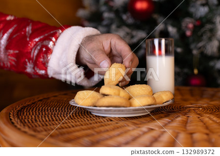 Santa Claus's hand is taking a cookie from a plate next to a glass of milk 132989372