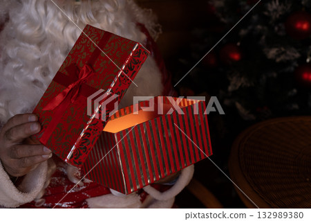 Santa Claus sits in a chair near a Christmas tree, holding a gift box Santa Claus sits in a chair near a Christmas tree, holding a gift box 132989380