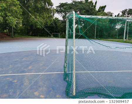 Outdoor futsal goalpost with striped metal frame and loose green net on blue sports court, standalone mini football goal in open ground surrounded by grass and trees, dedicated small-sided play area Outdoor futsal goalpost with striped metal frame and loose green net on blue sports court, standalone mini football goal in open ground surrounded by grass and trees, dedicated small-sided play area 132989457
