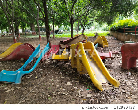 Broken and scattered plastic playground slides and ride-on equipment in park, yellow, red, blue pieces on muddy ground indicate neglect and disrepair Broken and scattered plastic playground slides and ride-on equipment in park, yellow, red, blue pieces on muddy ground indicate neglect and disrepair 132989458