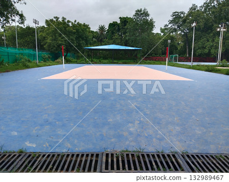 Outdoor court with blue flooring and peach playing area, empty sports ground featuring painted lines and net posts, surrounded by greenery and drainage channel in a quiet park environment 132989467