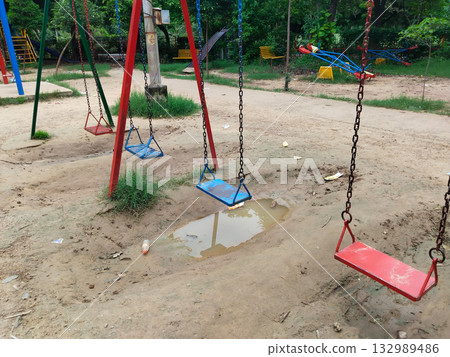 Blue and red metal swings with chains in outdoor playground, above muddy and uneven ground with puddles 132989486
