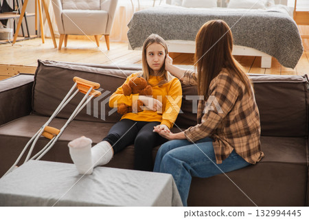 Girl with broken leg in cast sits with mother on couch at home. Sad little kid hugging teddy bear recovering from broken leg. Girl using cast crutches for quick rehabilitation. Mom supporting daughter Girl with broken leg in cast sits with mother on couch at home. Sad little kid hugging teddy bear recovering from broken leg. Girl using cast crutches for quick rehabilitation. Mom supporting daughter 132994445