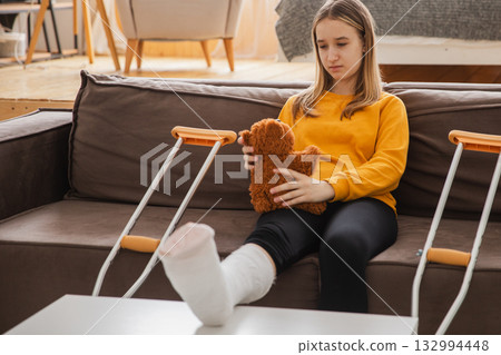 Young girl with broken leg cast sitting on sofa. Sad little kid hugging her teddy bear recovering from broken leg at home. Girl using cast and crutches. Accident injury fracture treatment foot bandage Young girl with broken leg cast sitting on sofa. Sad little kid hugging her teddy bear recovering from broken leg at home. Girl using cast and crutches. Accident injury fracture treatment foot bandage 132994448