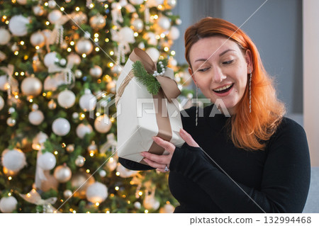 Merry Christmas. Woman unpacking open up gift box near Christmas tree. Girl in living room with Christmas tree holding opening gift box with surprise face. Woman celebrating Christmas eve at home Merry Christmas. Woman unpacking open up gift box near Christmas tree. Girl in living room with Christmas tree holding opening gift box with surprise face. Woman celebrating Christmas eve at home 132994468