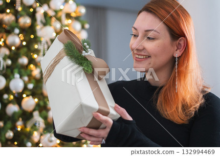 Merry Christmas. Woman unpacking open up gift box near Christmas tree. Girl in living room with Christmas tree holding opening gift box with surprise face. Woman celebrating Christmas eve at home 132994469