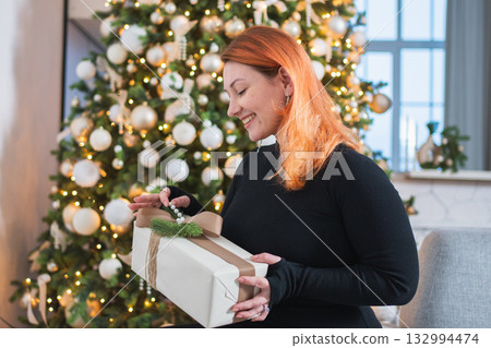 Merry Christmas. Woman unpacking open up gift box near Christmas tree. Girl in living room with Christmas tree holding opening gift box with surprise face. Woman celebrating Christmas eve at home 132994474
