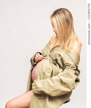 28 week, Elegant maternity portrait of pregnant woman in beige trench coat. Natural makeup and minimal studio light emphasize femininity and calm mood 28 week, Elegant maternity portrait of pregnant woman in beige trench coat. Natural makeup and minimal studio light emphasize femininity and calm mood 132994750