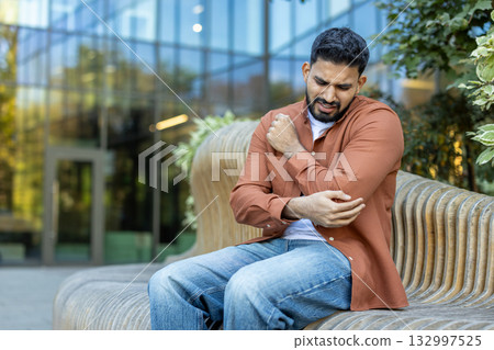 A man expresses pain by holding his elbow while sitting on a modern bench outdoors, surrounded by greenery and a glass building facade in the background. A man expresses pain by holding his elbow while sitting on a modern bench outdoors, surrounded by greenery and a glass building facade in the background. 132997525