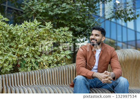 A cheerful young man in casual clothing sits on a wooden bench outdoors among lush greenery. The bright and modern urban environment enhances the warm and welcoming atmosphere. A cheerful young man in casual clothing sits on a wooden bench outdoors among lush greenery. The bright and modern urban environment enhances the warm and welcoming atmosphere. 132997534