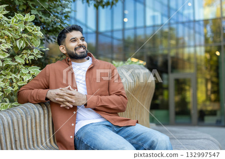 Young indian man smiling and looking away, sitting on a modern wooden bench outdoors with hands clasped, enjoying a moment of relaxation in an urban green space Young indian man smiling and looking away, sitting on a modern wooden bench outdoors with hands clasped, enjoying a moment of relaxation in an urban green space 132997547
