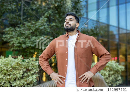 Confident indian manager taking a deep breath, enjoying fresh air in a green urban space, promoting mindfulness and work-life balance in a corporate setting 132997561