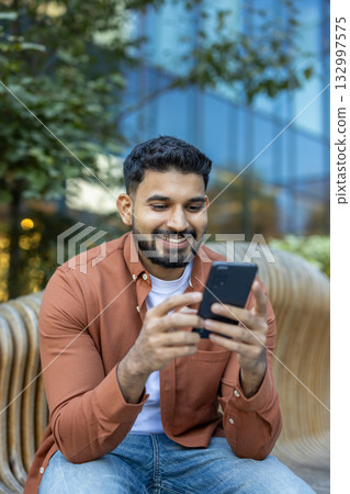 Young man holding and looking at a smartphone, smiling while sitting casually on a modern bench outdoors in a park setting with urban elements in the background, enjoying digital content 132997575