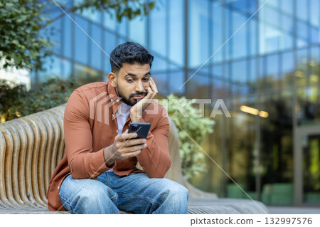 A man seated on an outdoor bench contemplating while looking at a smartphone. The setting includes modern architecture, natural greenery, and a calm environment that suggests contemplation 132997576
