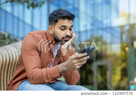 Young businessman sitting on a bench, looking bored while scrolling through his smartphone. Resting his face on his hand in front of a sleek, modern glass building 132997578