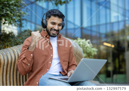 Happy indian man with headphones using a laptop on a park bench, celebrating success with a joyful fist pump while working or studying outdoors, smiling with confidence Happy indian man with headphones using a laptop on a park bench, celebrating success with a joyful fist pump while working or studying outdoors, smiling with confidence 132997579