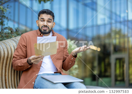 Young man sitting outdoors on a bench holding an open envelope with papers, looking confused and disappointed with the received news, reflecting on university failure Young man sitting outdoors on a bench holding an open envelope with papers, looking confused and disappointed with the received news, reflecting on university failure 132997583