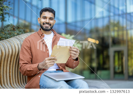Happy young man sitting outdoors on a modern bench, smiling and holding an open envelope with an acceptance letter, possibly receiving good news about his university admission 132997613