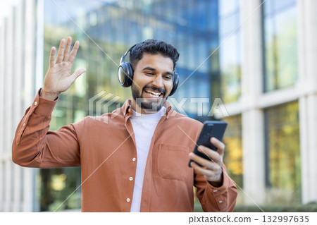 A cheerful person wearing headphones, interacting with a smartphone while outdoors, waving and smiling. A cheerful person wearing headphones, interacting with a smartphone while outdoors, waving and smiling. 132997635
