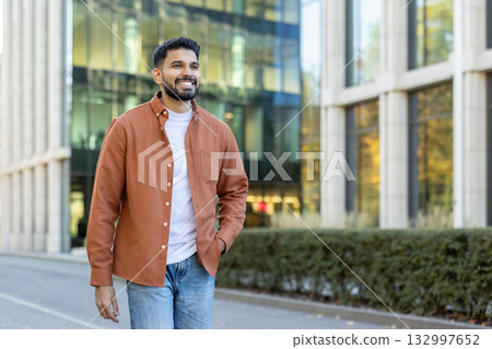 Young bearded indian businessman walking confidently near modern office buildings, smiling while looking away, enjoying the vibrant atmosphere of a successful career and future aspirations Young bearded indian businessman walking confidently near modern office buildings, smiling while looking away, enjoying the vibrant atmosphere of a successful career and future aspirations 132997652