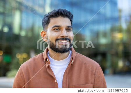 Smiling young indian businessman standing outside a modern office building, gazing away from the camera, exuding confidence and professionalism against a vibrant urban backdrop Smiling young indian businessman standing outside a modern office building, gazing away from the camera, exuding confidence and professionalism against a vibrant urban backdrop 132997655