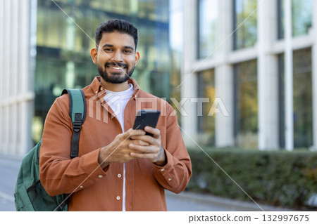 Young latin university student smiling while using a smartphone app near a campus building, dressed in casual clothes and carrying a green backpack, enjoying a vibrant day outdoors Young latin university student smiling while using a smartphone app near a campus building, dressed in casual clothes and carrying a green backpack, enjoying a vibrant day outdoors 132997675