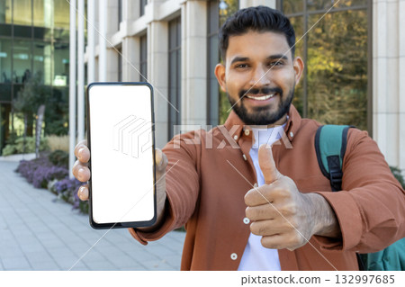 Happy young university student is showing a smartphone with a white screen and gesturing thumb up while standing outside a modern university building Happy young university student is showing a smartphone with a white screen and gesturing thumb up while standing outside a modern university building 132997685
