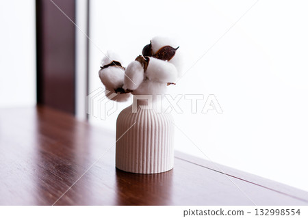 A white ceramic vase with a ribbed texture holds cotton branches. The background is bright and minimalistic, emphasizing the simplicity of the decor A white ceramic vase with a ribbed texture holds cotton branches. The background is bright and minimalistic, emphasizing the simplicity of the decor 132998554