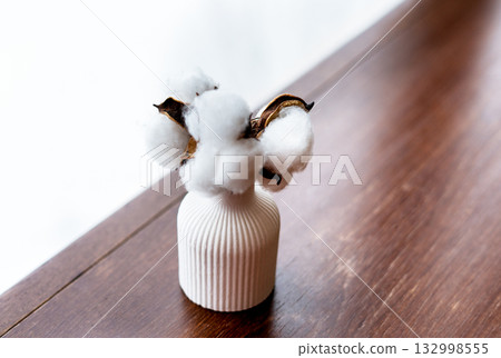A white ceramic vase holds cotton branches with fluffy white bolls. The vase is placed on a wooden surface, creating a minimalist decor scene. A white ceramic vase holds cotton branches with fluffy white bolls. The vase is placed on a wooden surface, creating a minimalist decor scene. 132998555