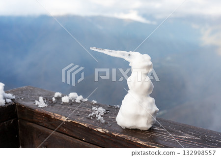 small snowman made of snow and an icicle for a nose sits on a wooden surface. In the background are a cloudy sky and distant mountains, winter landscape small snowman made of snow and an icicle for a nose sits on a wooden surface. In the background are a cloudy sky and distant mountains, winter landscape 132998557