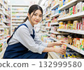 A female convenience store clerk with a ponytail and an apron arranging products 132999580