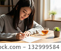 A long-haired woman wearing a gray knit sweater and writing a letter seriously in warm light 132999584