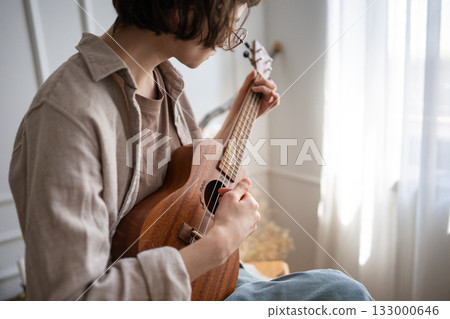 Young woman rehearsing ukulele chords at home, refining finger placement and strumming technique 133000646
