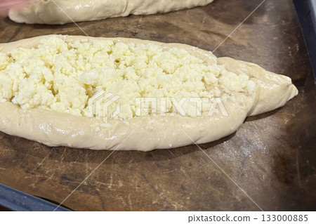Dough shaped like a boat on a baking tray before baking, topped with shredded suluguni cheese, preparing a Georgian cuisine dish, khachapuri, close-up 133000885