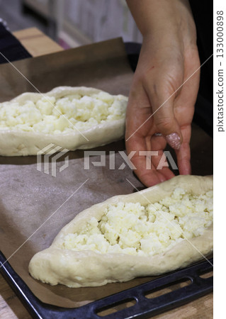 Prepared dough pieces on a baking tray, dough shaped into boats filled with shredded suluguni cheese, woman preparing a Georgian cuisine dish, khachapuri. Prepared dough pieces on a baking tray, dough shaped into boats filled with shredded suluguni cheese, woman preparing a Georgian cuisine dish, khachapuri. 133000898