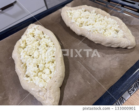 Prepared dough pieces on a baking tray, dough shaped into boats filled with shredded suluguni cheese, woman preparing a Georgian cuisine dish, khachapuri. Prepared dough pieces on a baking tray, dough shaped into boats filled with shredded suluguni cheese, woman preparing a Georgian cuisine dish, khachapuri. 133000901