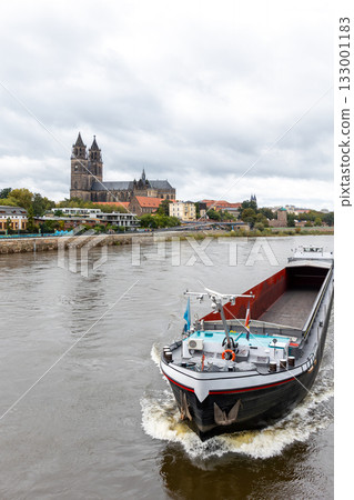 Cargo barge ship sails river Elbe historic Magdeburg city skyline cathedral Dom towers and colorful autumn trees. Overcast sky calm water create tranquil atmosphere European river inland transport 133001183