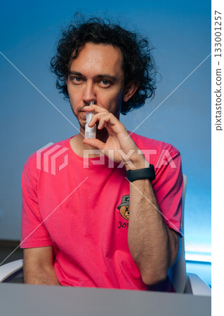 Young man with curly hair wearing pink t-shirt using nasal spray to alleviate allergy symptoms or congestion, sitting at a desk on isolated vibrant blue background. Concept of healthy lifestyle. 133001257
