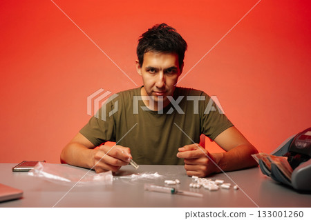 Portrait of serious man preparing to snort cocaine with rolled banknote sitting at table with pills and syringe on red background, looking at camera, representing drug addiction and substance abuse. 133001260