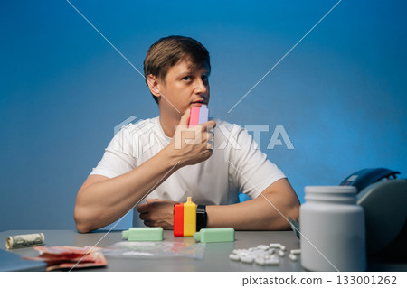 Electronic cigarette dealer displaying multiple colorful vaping devices amid smoke, surrounded by pills, cash and plastic bags on blue tabletop surface, studio shot portrait on isolated background.. 133001262