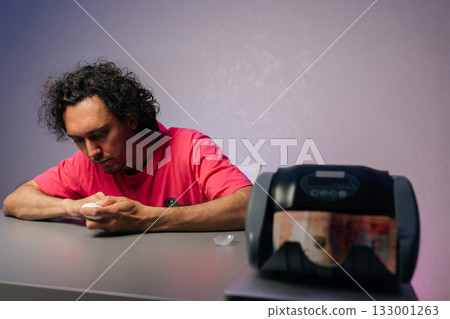 Male entrepreneur grooming fingernails using electric clipper while counting money with electronic counting machine, combining personal care and financial management at home workspace. 133001263