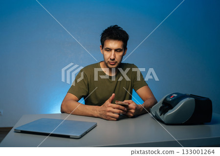 Pensive bank employee male counting currency using automatic banknote counter machine at modern dark workplace, sitting at desk with laptop in room illuminated by soft blue light in background. 133001264
