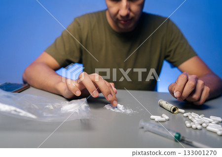 Unrecognizable drug user preparing dose of cocaine or heroin using rolled-up dollar bill on table with pills and plastic baggies, illustrating drug abuse and addiction. Concept of substance abuse. Unrecognizable drug user preparing dose of cocaine or heroin using rolled-up dollar bill on table with pills and plastic baggies, illustrating drug abuse and addiction. Concept of substance abuse. 133001270