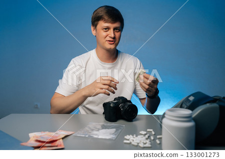 Young photographer preparing drugs with rolled banknote at studio, sitting at desk with camera, struggling with addiction, studio shot portrait on isolated background. Concept of substance abuse. 133001273
