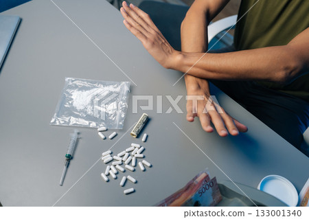 Recovering drug user pushing away narcotics, surrounded by scattered medical tools and paraphernalia, representing personal battle against substance dependency. Concept of healthy lifestyle. 133001340