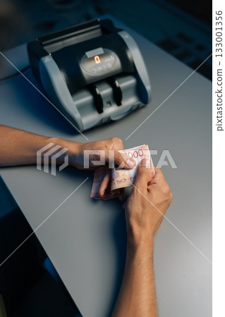 Close-up top view of cashier counting Russian rubles banknotes late at night after closing store using money counting machine in background. Concept of financial accounting in modern office. Close-up top view of cashier counting Russian rubles banknotes late at night after closing store using money counting machine in background. Concept of financial accounting in modern office. 133001356