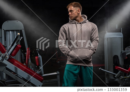 athletic person stands in the gym wearing a gray hoodie embodying confidence and strength while surrounded by exercise equipment. The lighting highlights their focused demeanor. athletic person stands in the gym wearing a gray hoodie embodying confidence and strength while surrounded by exercise equipment. The lighting highlights their focused demeanor. 133001359