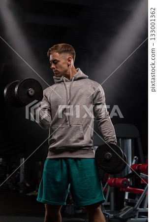 An athlete in a gray hoodie is focused on doing bicep curls with dumbbells. The gym setting is equipped with various workout machines. Evening light creates a vibrant atmosphere. An athlete in a gray hoodie is focused on doing bicep curls with dumbbells. The gym setting is equipped with various workout machines. Evening light creates a vibrant atmosphere. 133001362